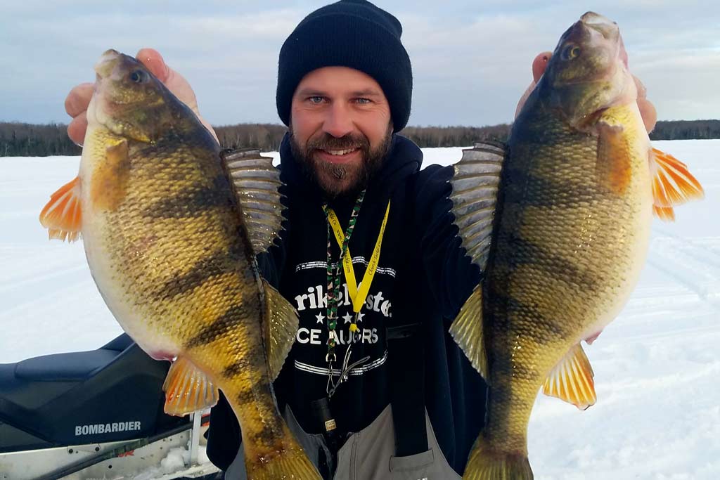 A smiling villous man in a hat holding two Yellow Perch, with frozen waters overdue him