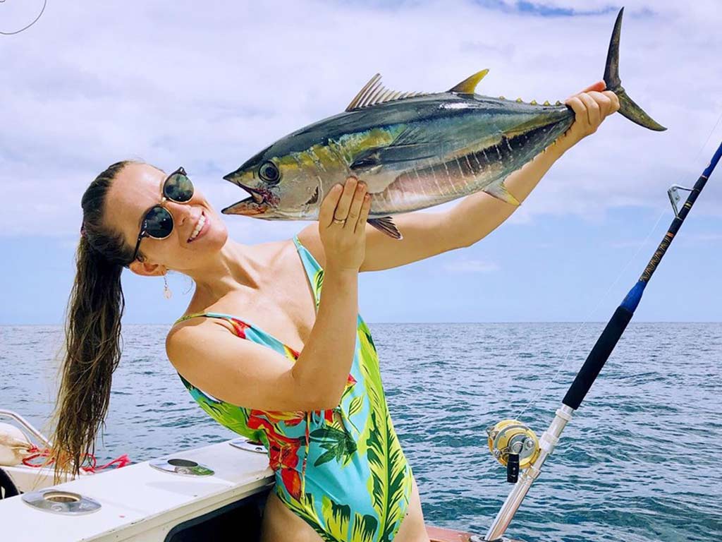 A woman posing for a photo, holding a Yellowfin Tuna she caught high up in the air.