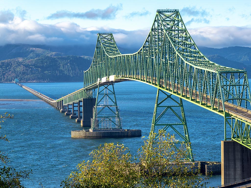 Astoria Bridge on a warm summer day, with some foliage in the foreground and mountains in the background.