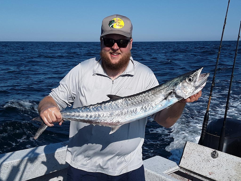 A smiling angler stands at the back of a boat, holding a King Mackerel he recently caught.