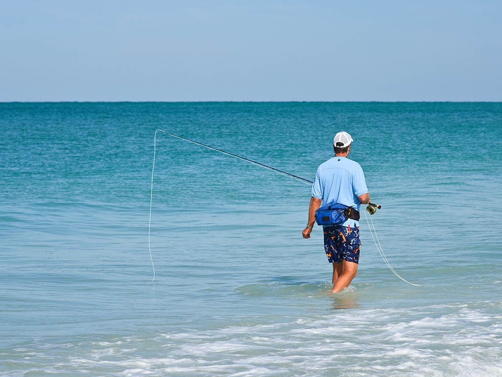 A man in a blue shirt wades in the surf in Destin, FL, while holding a fly fishing rod