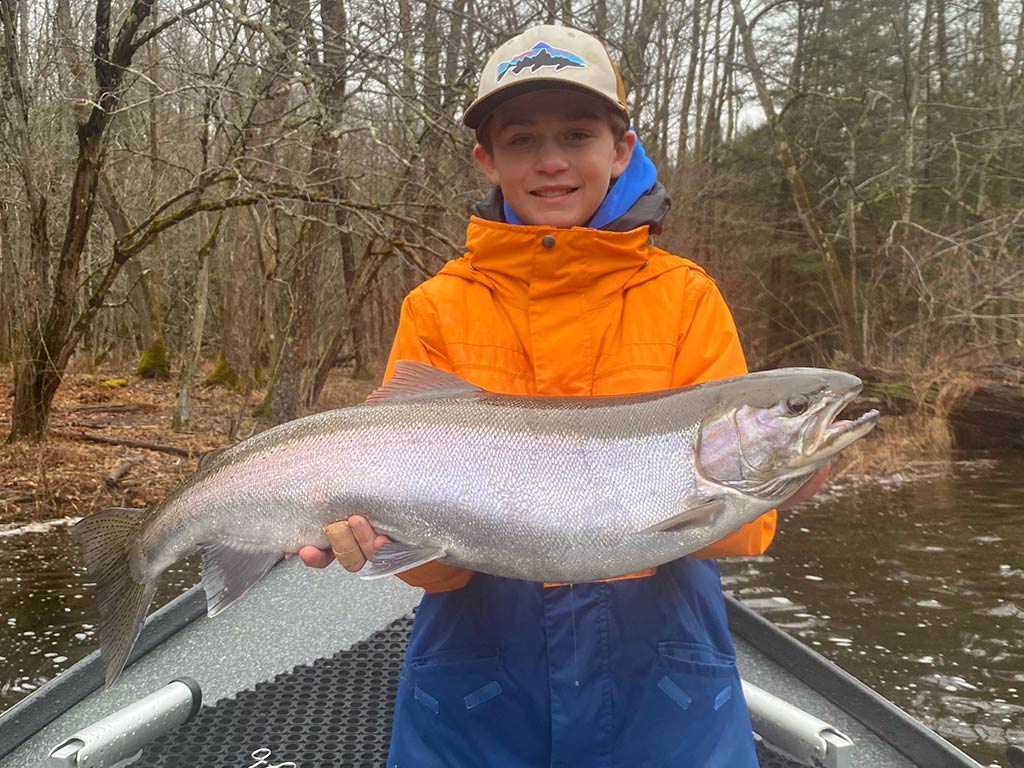 A young angler in a winter coat and cap holding a large Steelhead caught in the Salmon River