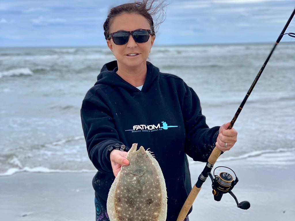 An angler stands on a beach holding a fishing rod and a Flounder she just caught.