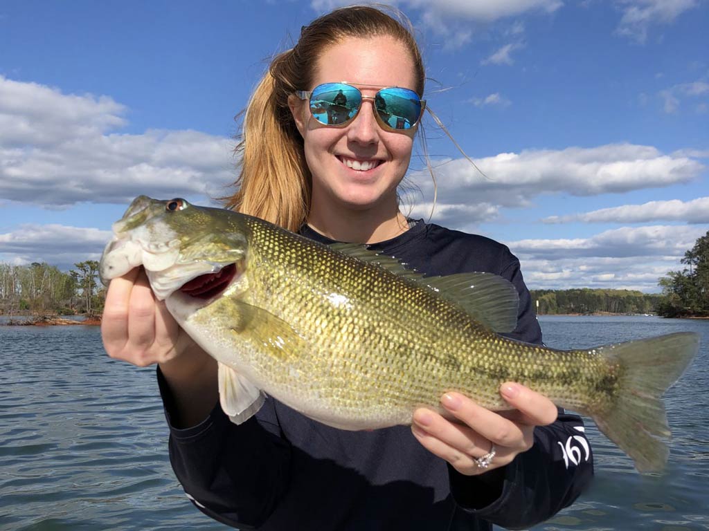 A young woman holding a Bass with both her hands while standing on a South Carolina fishing charter boat on a sunny spring day