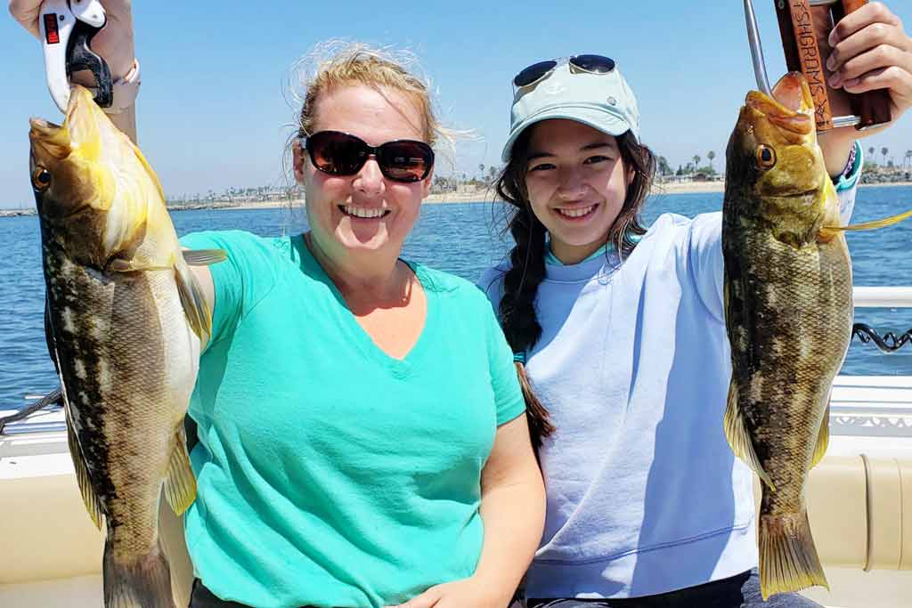 A mother and daughter sitting on a fishing boat, each holding a big Calico Bass with a beach behind them in the distance