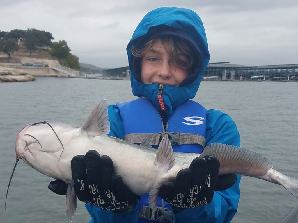 A photo of a kid wearing gloves and warm jacket while holding a Catfish and standing on a charter boat on a cold day