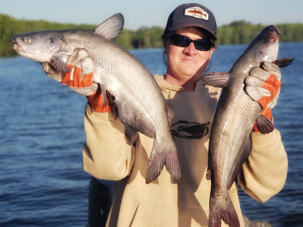 A n angler standing on a boat and proudly holding two Catfish – one in each hand