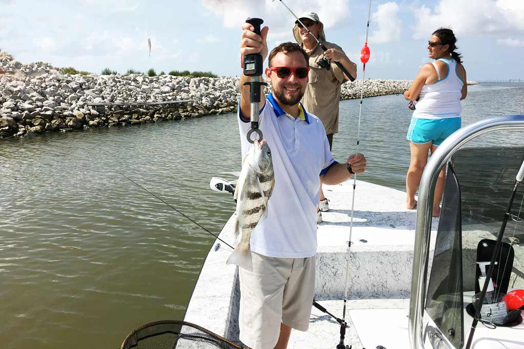 A smiling man in sunglasses holding a Black Drum fish, standing on a boat, with two women behind him, and water in the background