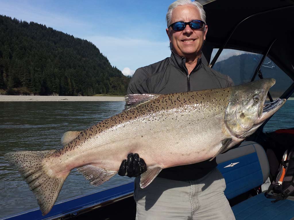 An angler on a boat, holding a big Chinook Salmon he caught fishing on the Fraser River, with the water and greenery behind him.