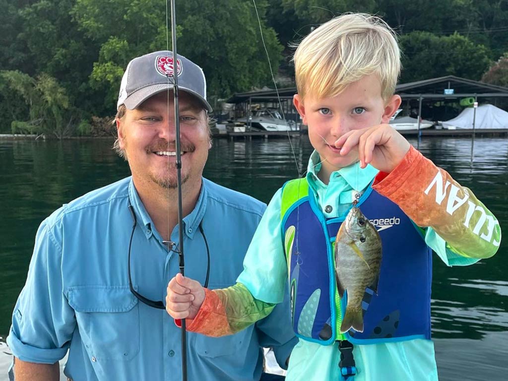 A cute photo of a child wearing a life jacket while holding a Crappie in one hand and a fishing rod in other, while standing on a charter boat and posing next to an adult angler