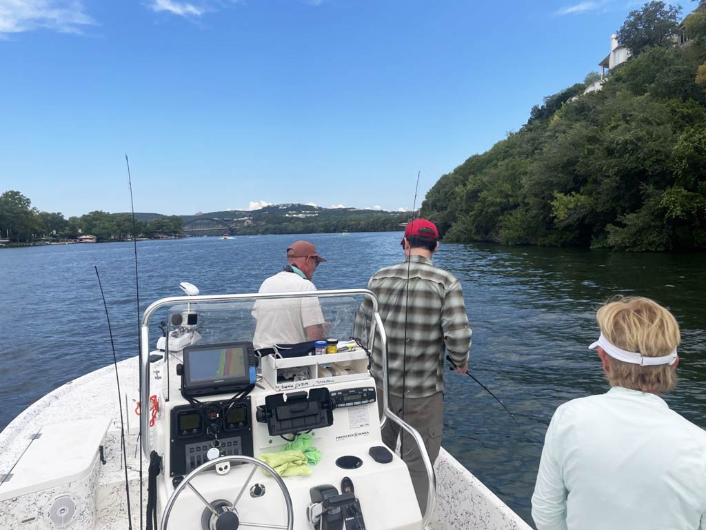 A photo of three anglers standing on a charter boat and angling on the Colorado River during a bright day surrounded by greenery