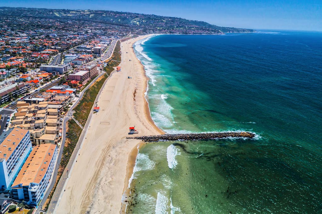 An aerial view of the Redondo Beach jetty and sandy beach and blue waters surrounding it on the right of the image