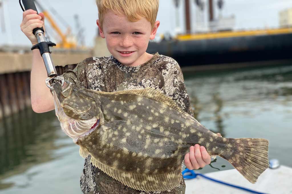 A small boy in a camouflage shirt holding a Flounder, with water in the background