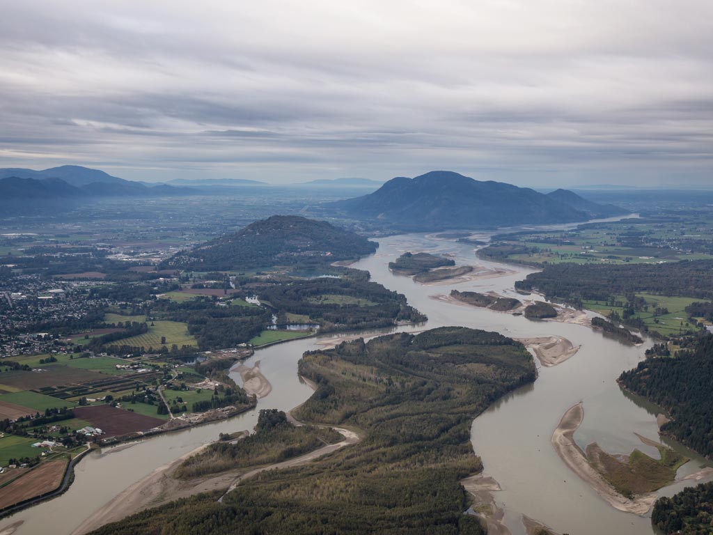 An aerial photo of the Fraser River near Chilliwack, taken on a cloudy day, with the river winding around some green islands, with mountains in the distance.