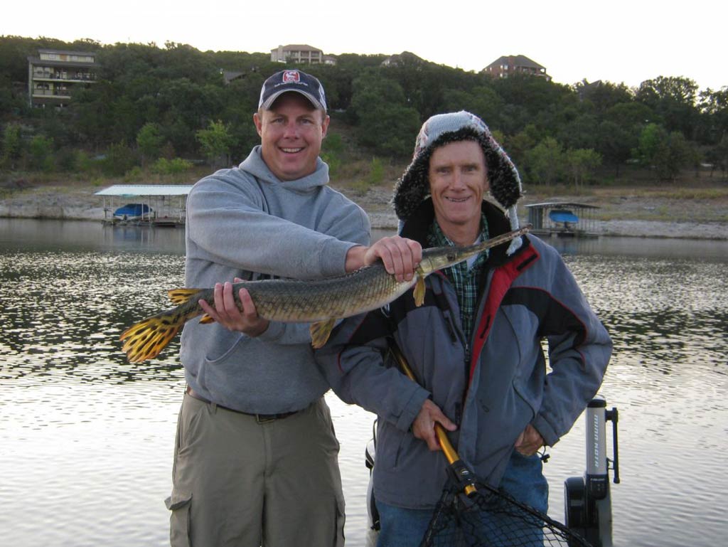 A photo of two satisfied anglers standing on a charter fishing boat while one angler holds a net and the other is showing off a Gar