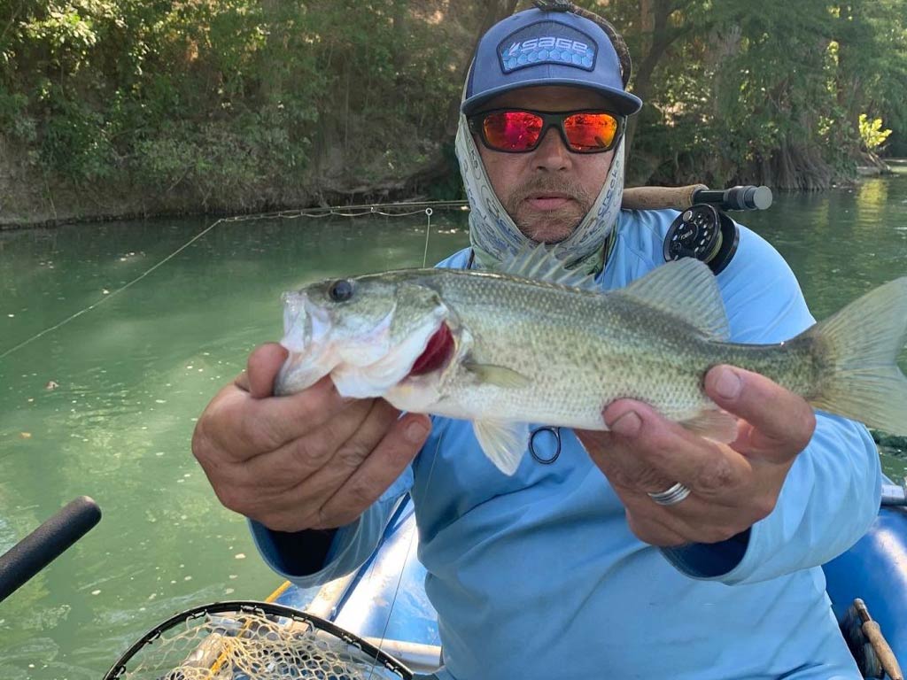 An angler wearing a cap and pair of sunglasses while sitting in a kayak and showing the fish he caught while angling in Austin, with a fly fishing rod over his shoulder