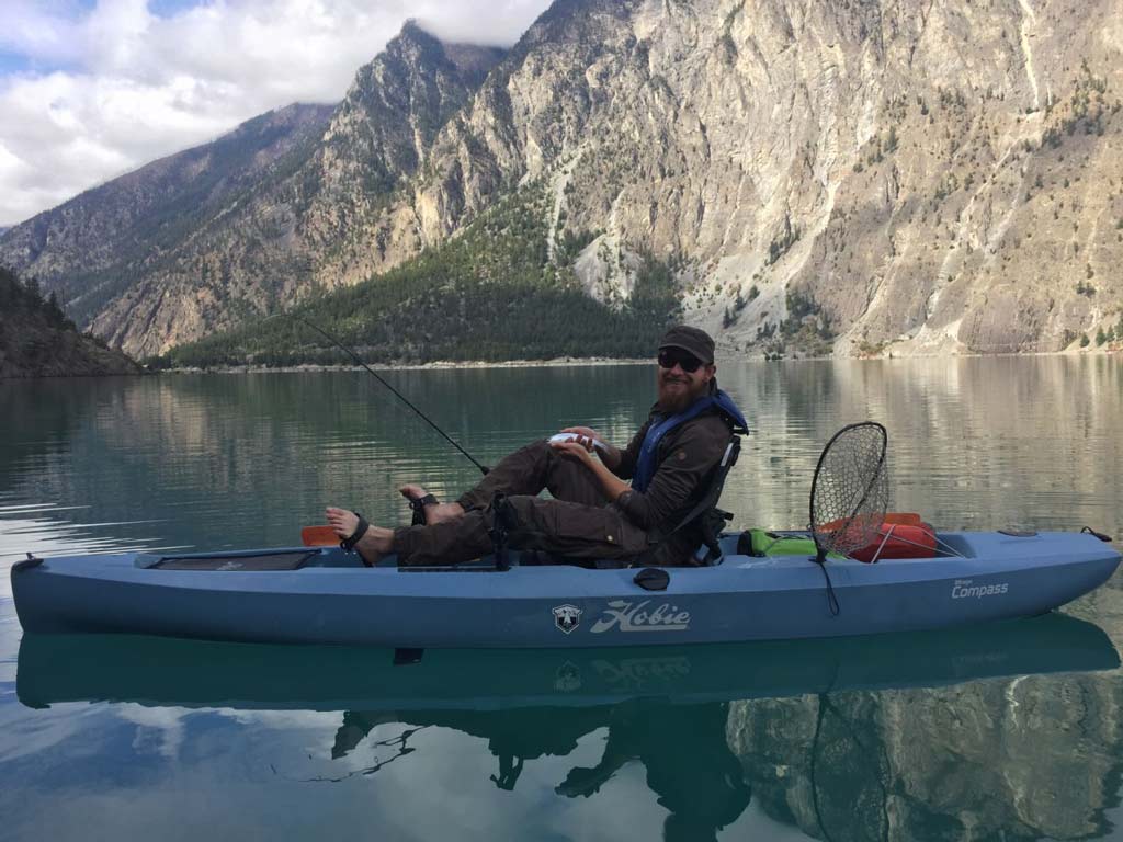 A smiling kayak angler holding a small fish he caught, late in the day on the calm waters of British Columbia.