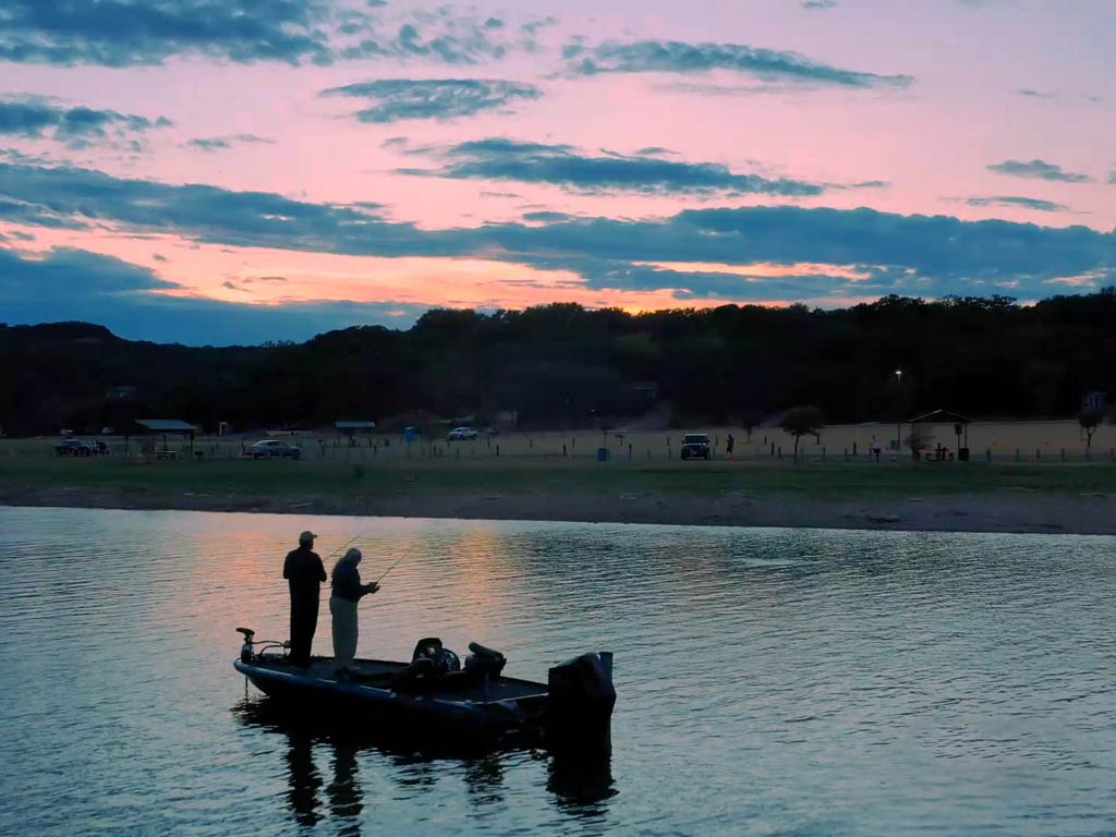 A sunset view of two anglers standing on a charter boat near the shore and fishing on Lake Travis