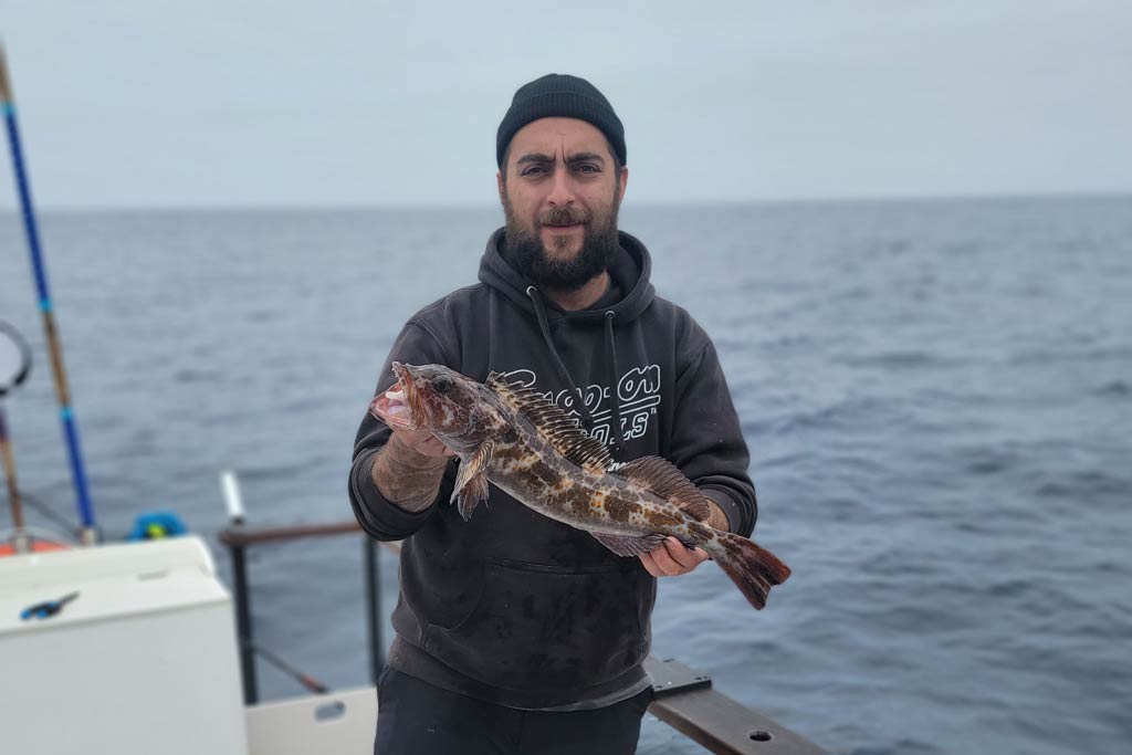 A bearded man in a cap standing on a fishing boat, holding a small Lingcod, with water and grey skies behind him