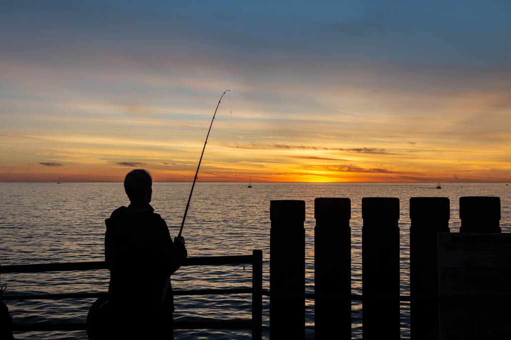 A sihouette of a fisherman holding a fishing rod with his line in the water, standing on the Redondo Beach Pier after sunset