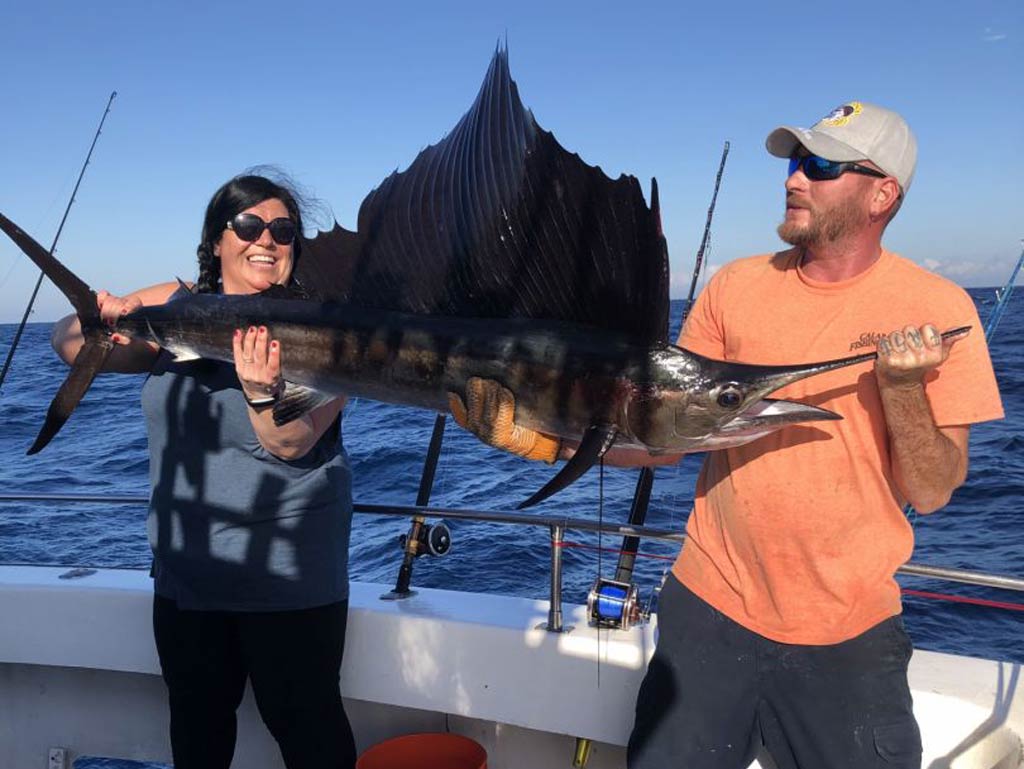A man and a woman standing on a fishing charter boat and holding a big Sailfish