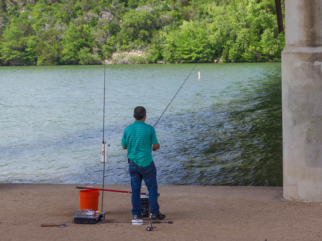 A photo of an angler taken from behind while he's standing on the shore and fishing in a river under a bridge 