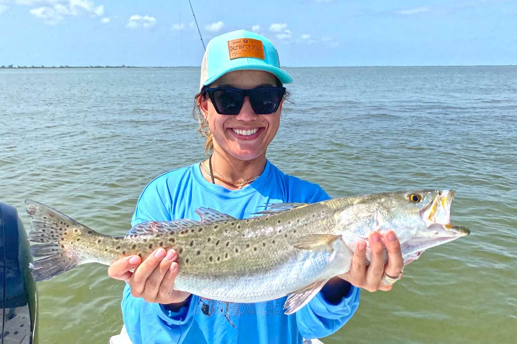 A smiling woman in a cap and sunglasses holding a big Speckled Trout, with clear skies and greenish waters behind her