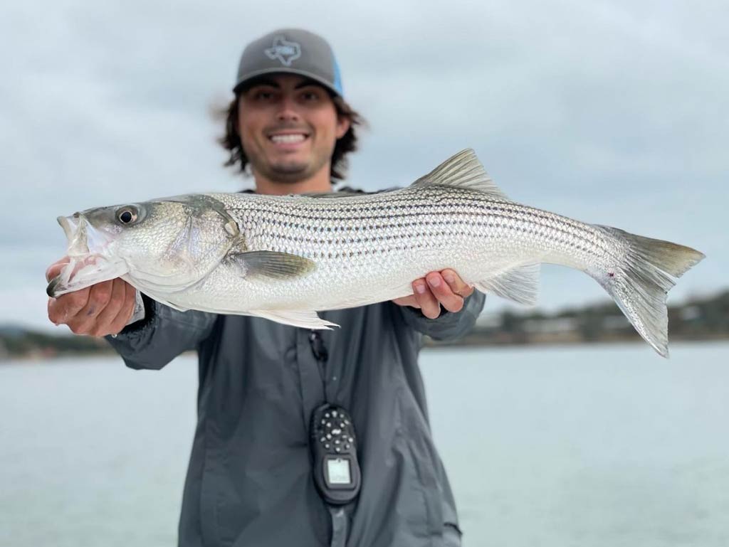 An angler proudly showing off his Striped Bass catch with both hands while standing on a charter boat against a blurred background