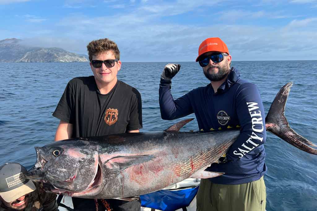 Two men standing on a fishing boat, holding a big Bluefin Tuna they caught, with open waters and clear skies in the background