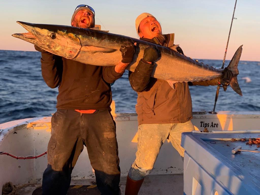 A sunset photo of two anglers on a boat at sunset, holding a big Wahoo while shouting happily