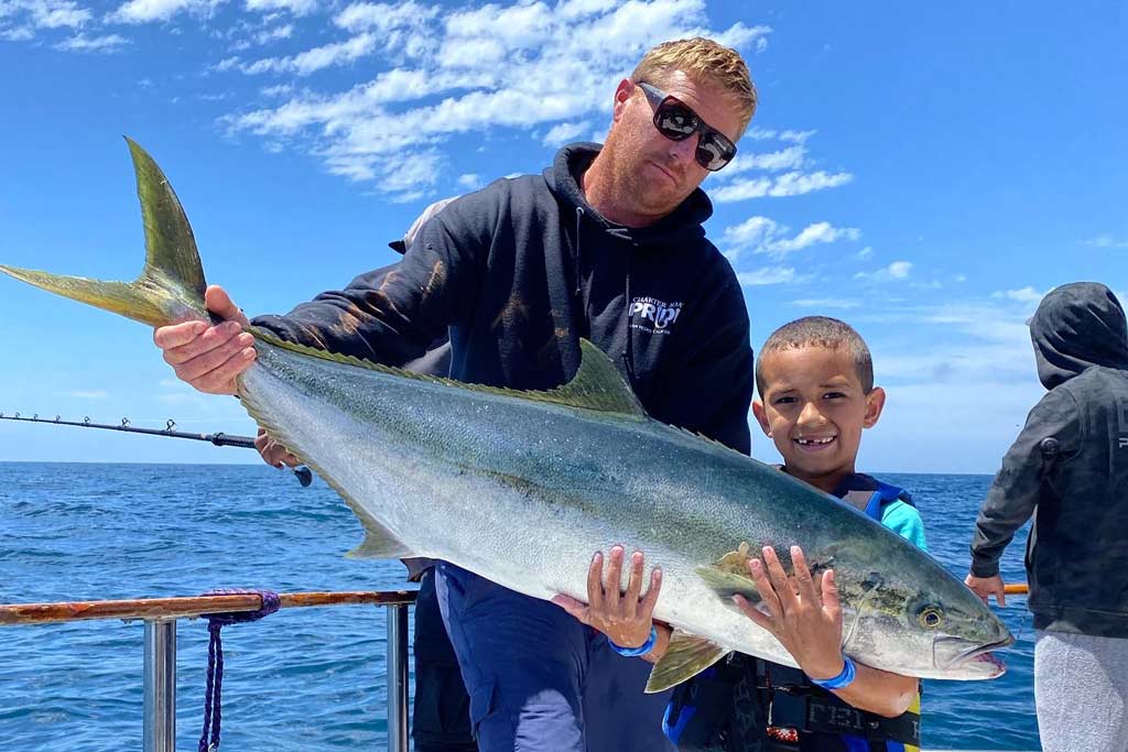 A man and a child, standing on a charter fishing boat, together holding a Yellowtail Amberjack they caught, with water and an angler casting a line behind them