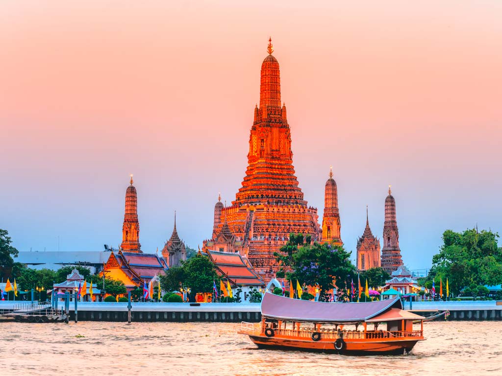 A sunset view of the Wat Arun Buddhist temple in Bangkok from the river, with a traditional Thai boat visible in the forefront at sunset.