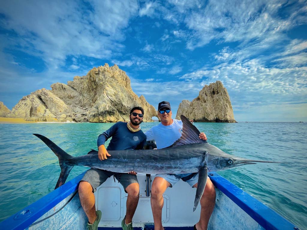 An excellent shot of two anglers sitting on a Cabo San Lucas fishing boat and holding a big Marlin with both hands while posing against the famous arch in the background on a sunny spring day
