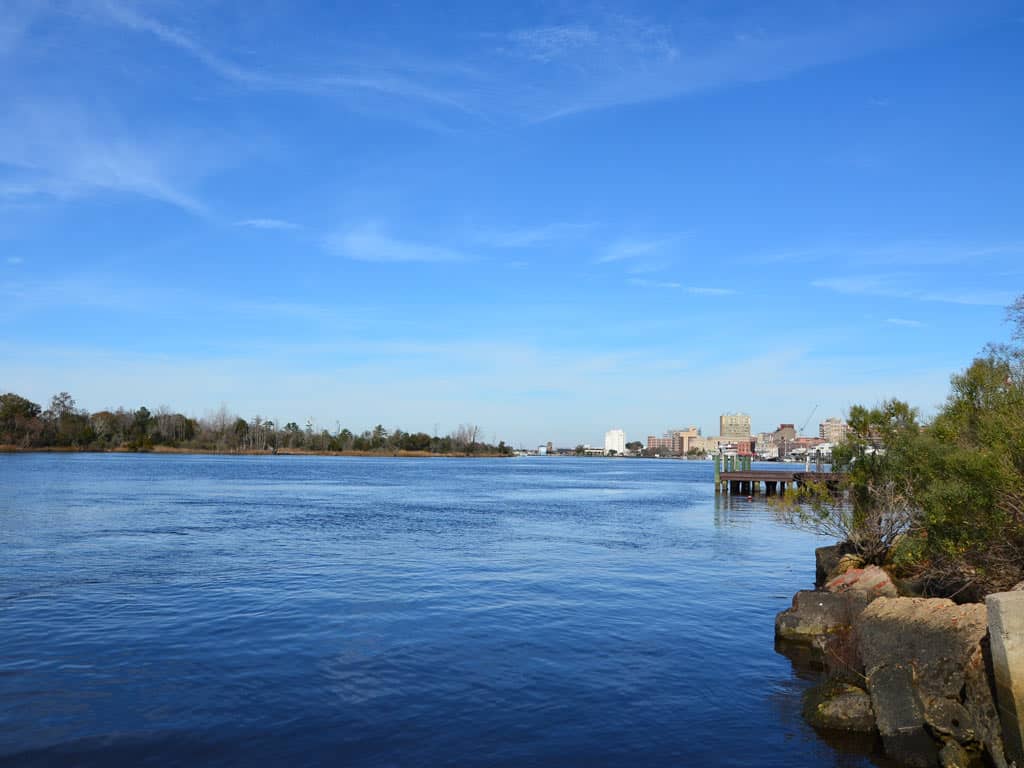 A view low-angle shot of the Cape Fear River near Wilmington, NC, with a rocky shoreline visible on the right of the image, along with a city in the distance across the water.