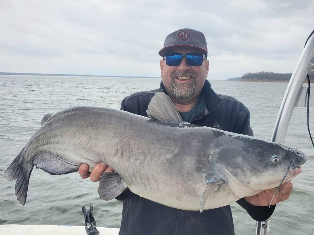 A photo of an angler wearing a cap and a pair of sunglasses while standing on a boat and proudly showing off a big Catfish caught while fishing the Red River on a cloudy fall day