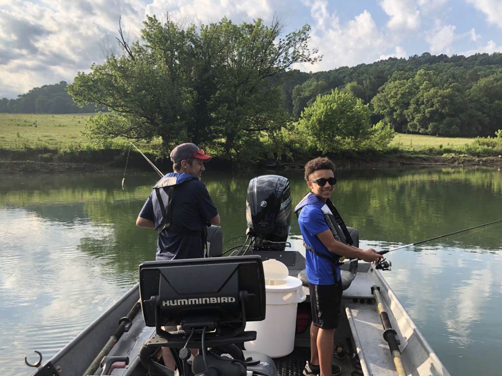 A photo of two youthful anglers standing on the back of a Tennessee charter fishing boat and casting a line on a bright spring day, with a green shoreline visible in the background