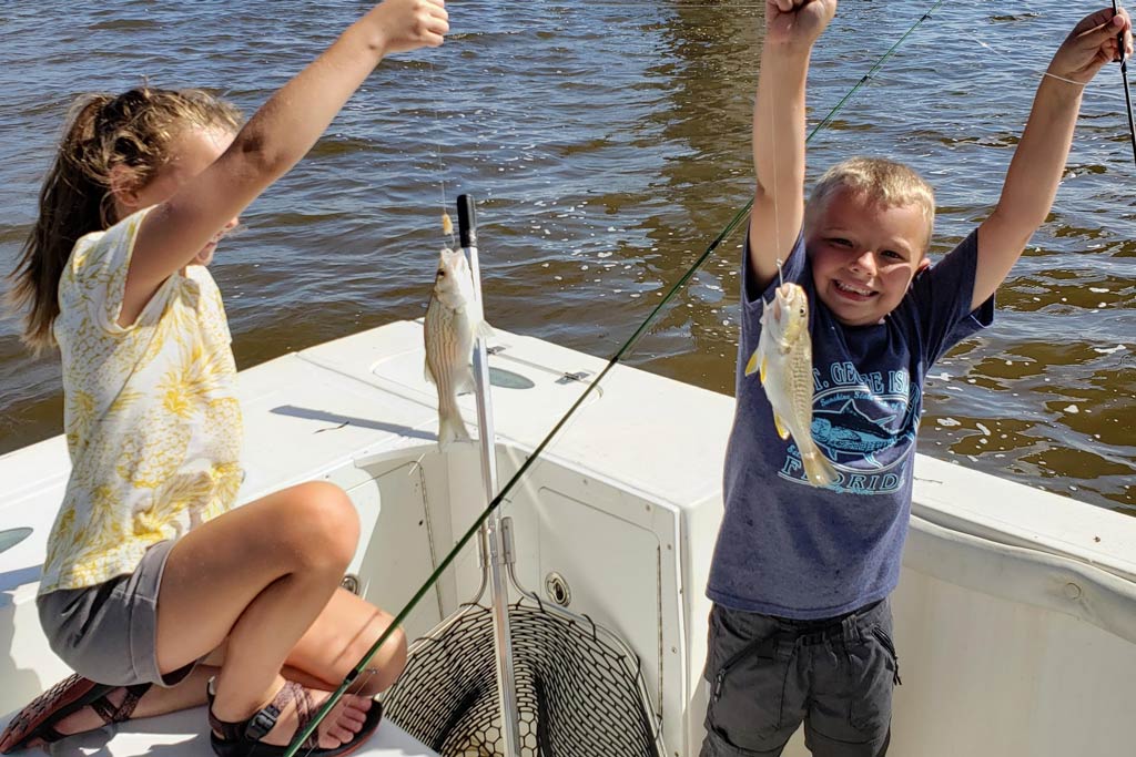 Two kids standing on a fishing boat, each holding a small fish they caught on their fishing lines, with brown waters behind them