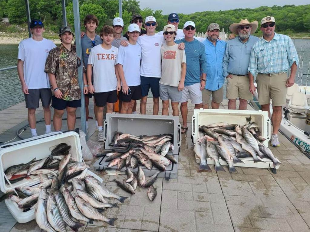 A group photo of fourteen happy anglers standing on the dock while posing behind three coolers filled with fish they caught during their Red River fishing trip on a spring day