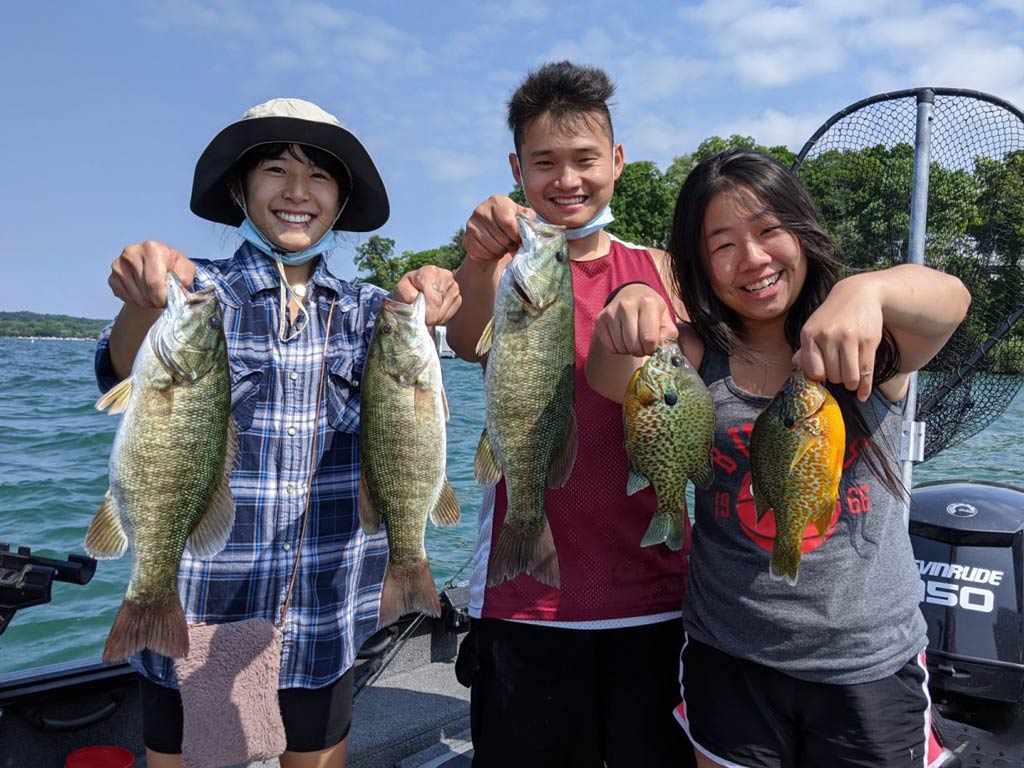 A photo of three young anglers standing on a charter boat and posing while holding some Bass and Sunfish against greenery and the shoreline in the background