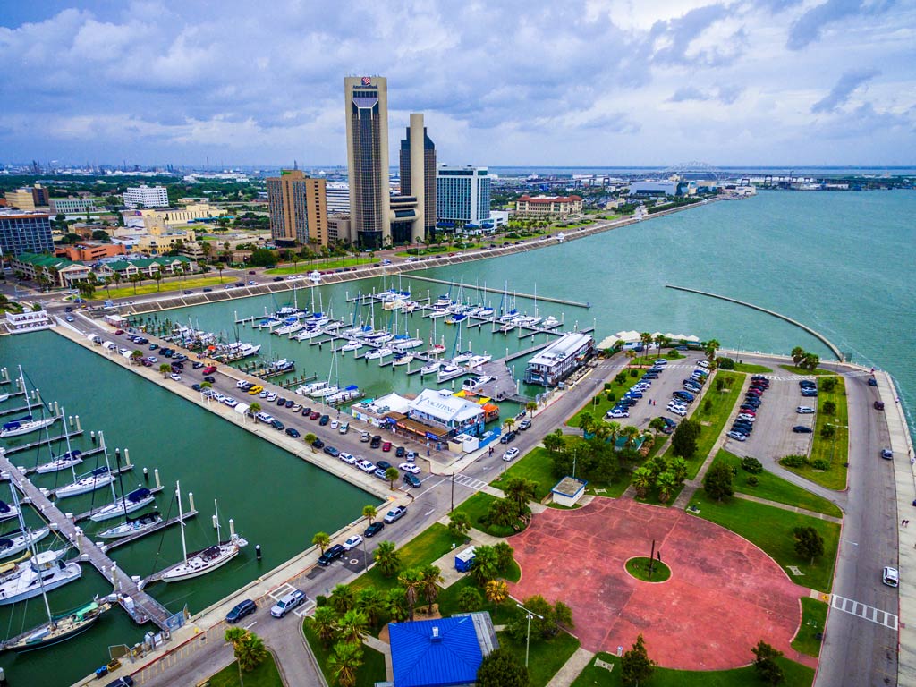 An aerial view of Corpus Christi’s waters, a marina, and boats, with the city's skyline in the background on a cloudy day