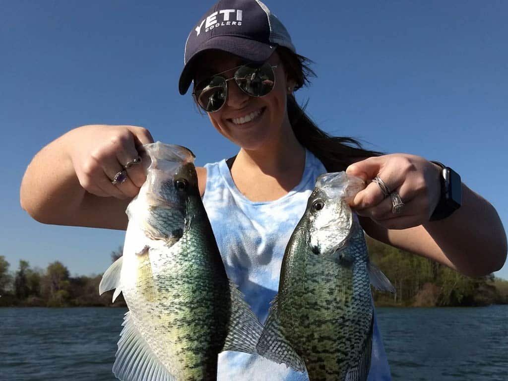 A photo of a female angler wearing a cap and sunglasses standing on a charter fishing boat and holding a Crappie in each hand on a sunny day 