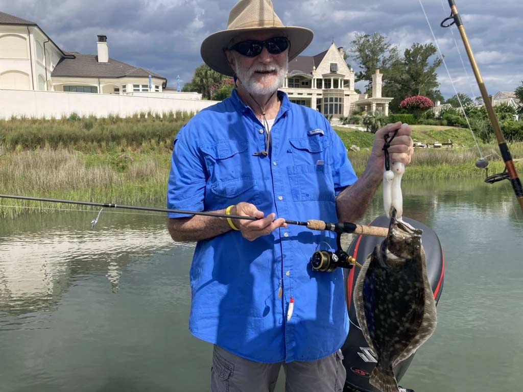 An angler holding a fishing rod in his right hand and a Flounder he caught in his left hand, while standing on a small boat in marshland, with a house facing the water behind him.