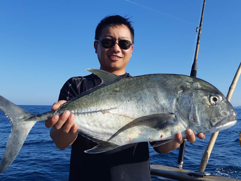 A smiling angler in sunglasses holding a Giant Trevally he caught fishing in Thailand, with two fishing rods, the ocean, and clear skies visible behind him.