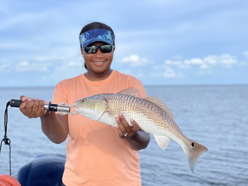 Happy black female angler in a visor, sunglasses, and orange t-shirt holding a redfish while standing on a boat.