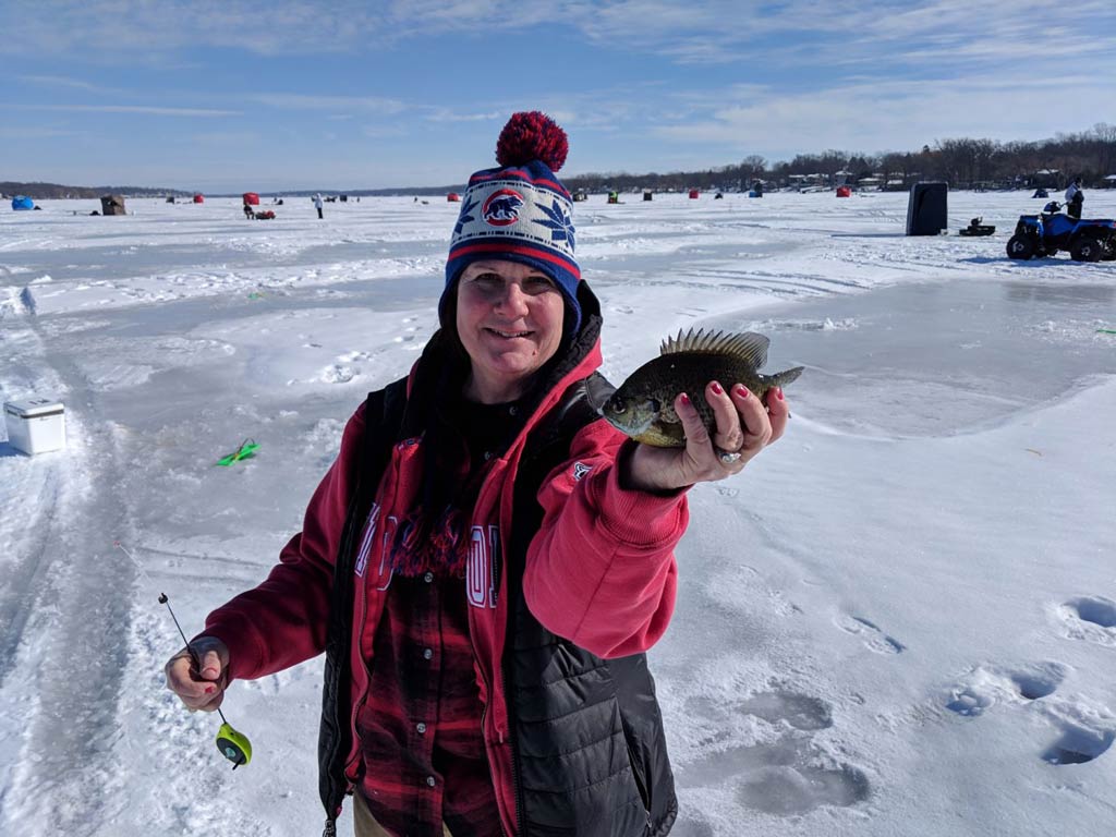 A photo of an angler wearing winter clothes and standing on a snow-covered lake while holding a small fish in one hand