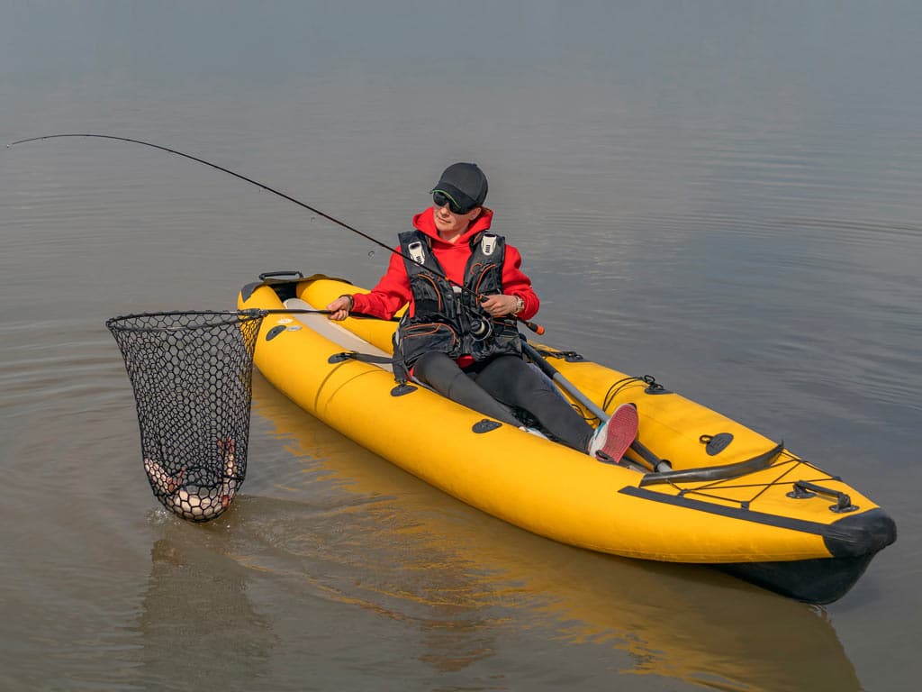 A female angler in a yellow kayak, netting a Pike, while holding a fishing rod in her other hand.