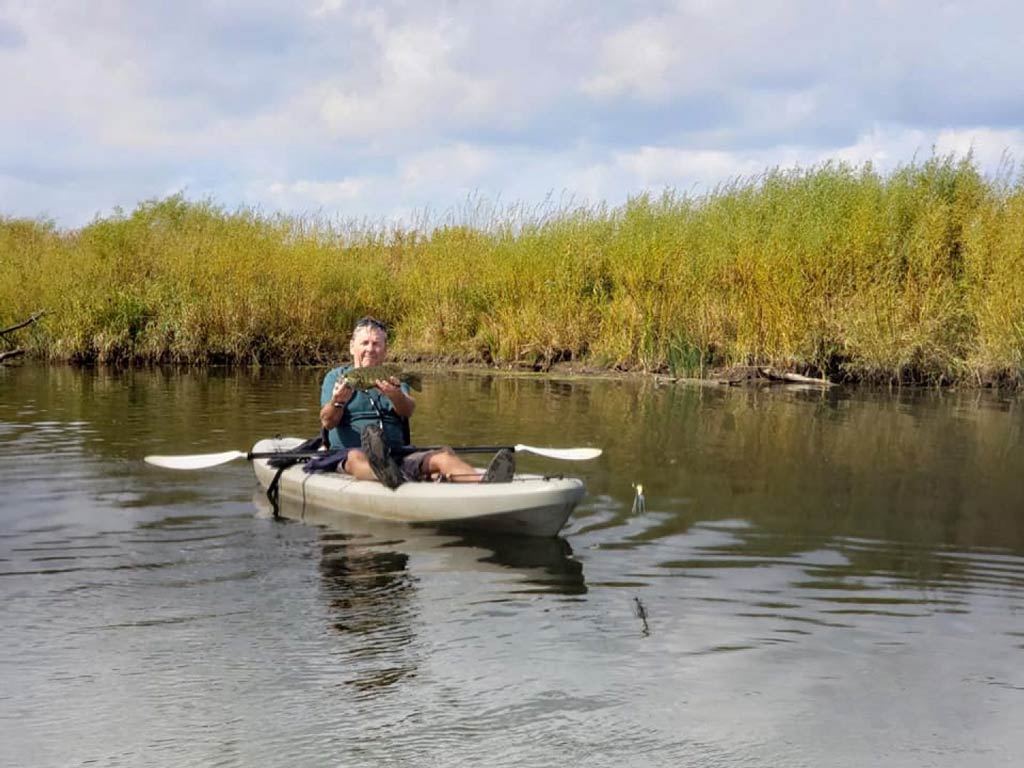 A photo of an angler sitting in a kayak and showing off a fish caught while kayak fishing on a river, with a grassy riverbank behind him