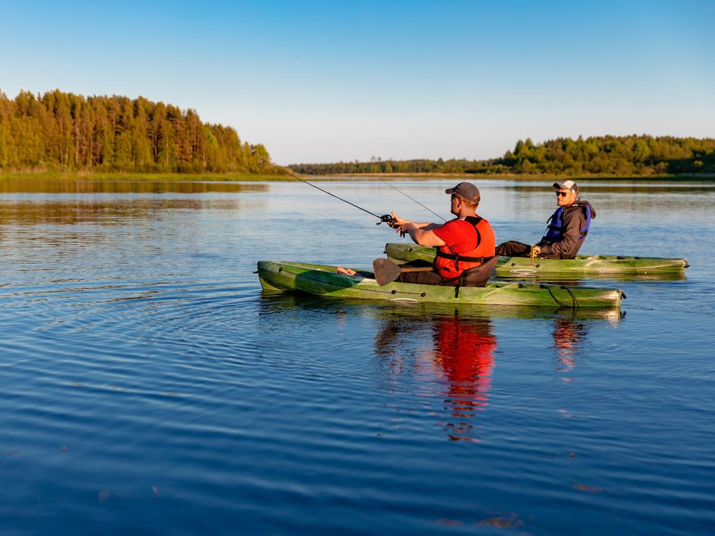 A photo of two anglers sitting in their kayaks near the shoreline and kayak angling on the river on a bright and sunny spring day