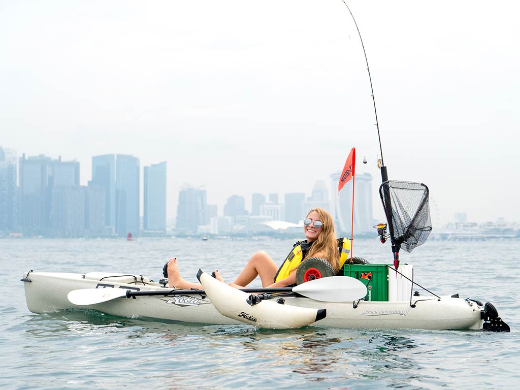 A view across the water towards a female sitting in a fishing kayak, with Singapore's skyline behind her on a cloudy day