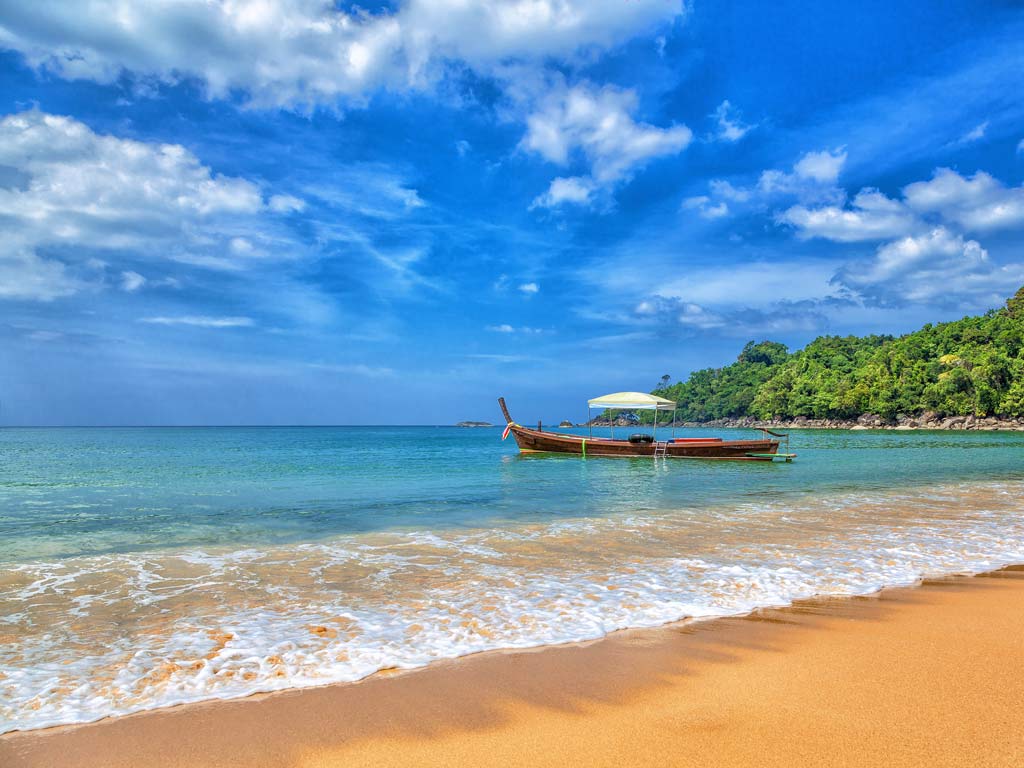 A photo of a beautiful beach in Khao Lak and a Thai longboat in the water, with calm ocean waters and greenery visible in the background.